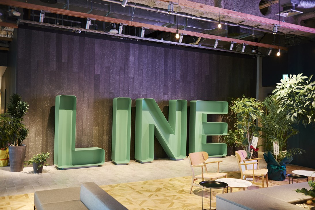 Large green LINE logo letters in the lobby of Line's Tokyo offices, with potted plants and wooden chairs nearby.