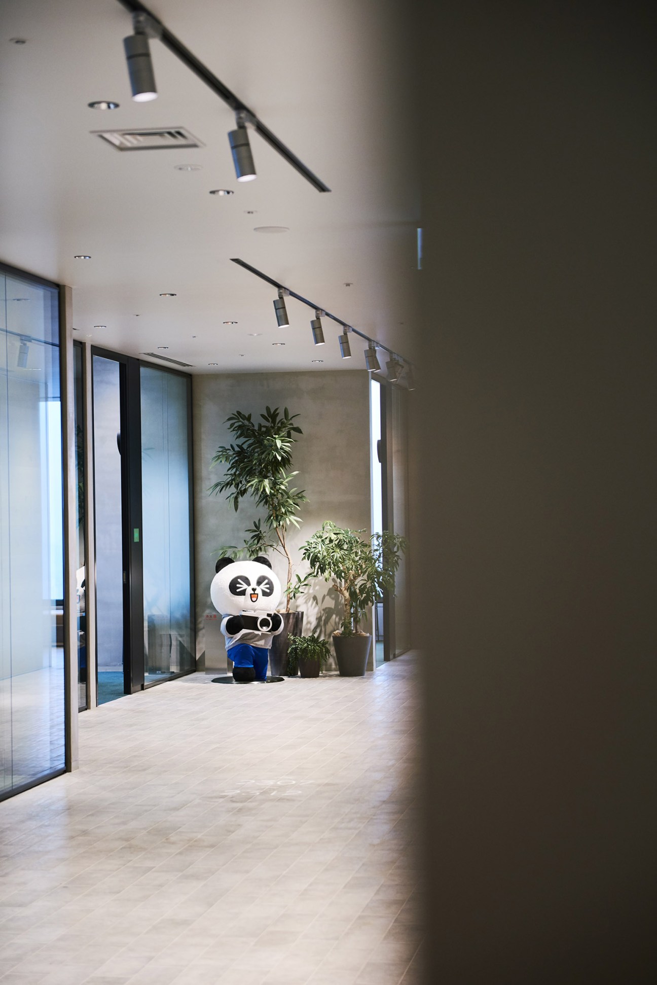A life-sized white panda character figure with black markings standing in a modern hallway with potted plants.