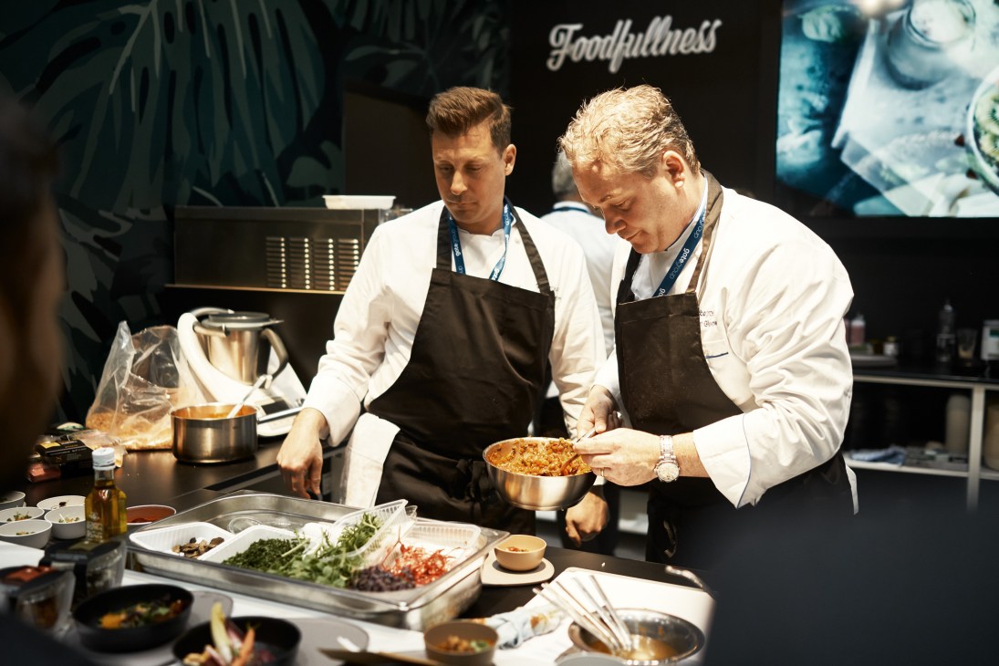Two chefs in white chef coats and black aprons prepare food at a cooking station during an airline catering expo demonstration.