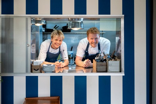Two chefs in white uniforms and aprons smiling through a kitchen service window