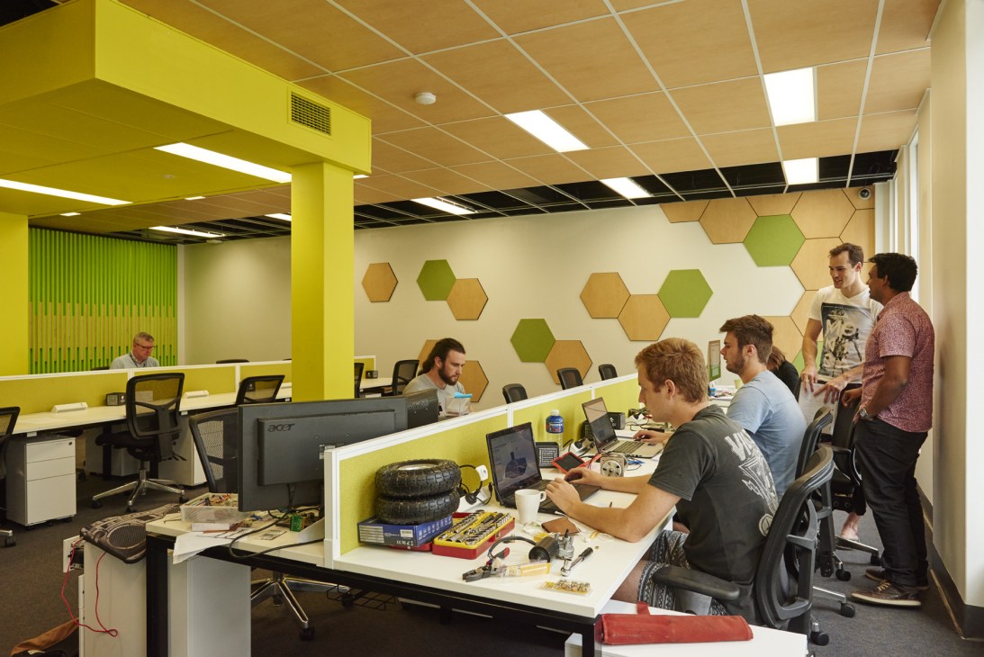 A modern co-working space with bright yellow structural elements, hexagonal wall panels, and professionals working at desks with computers.
