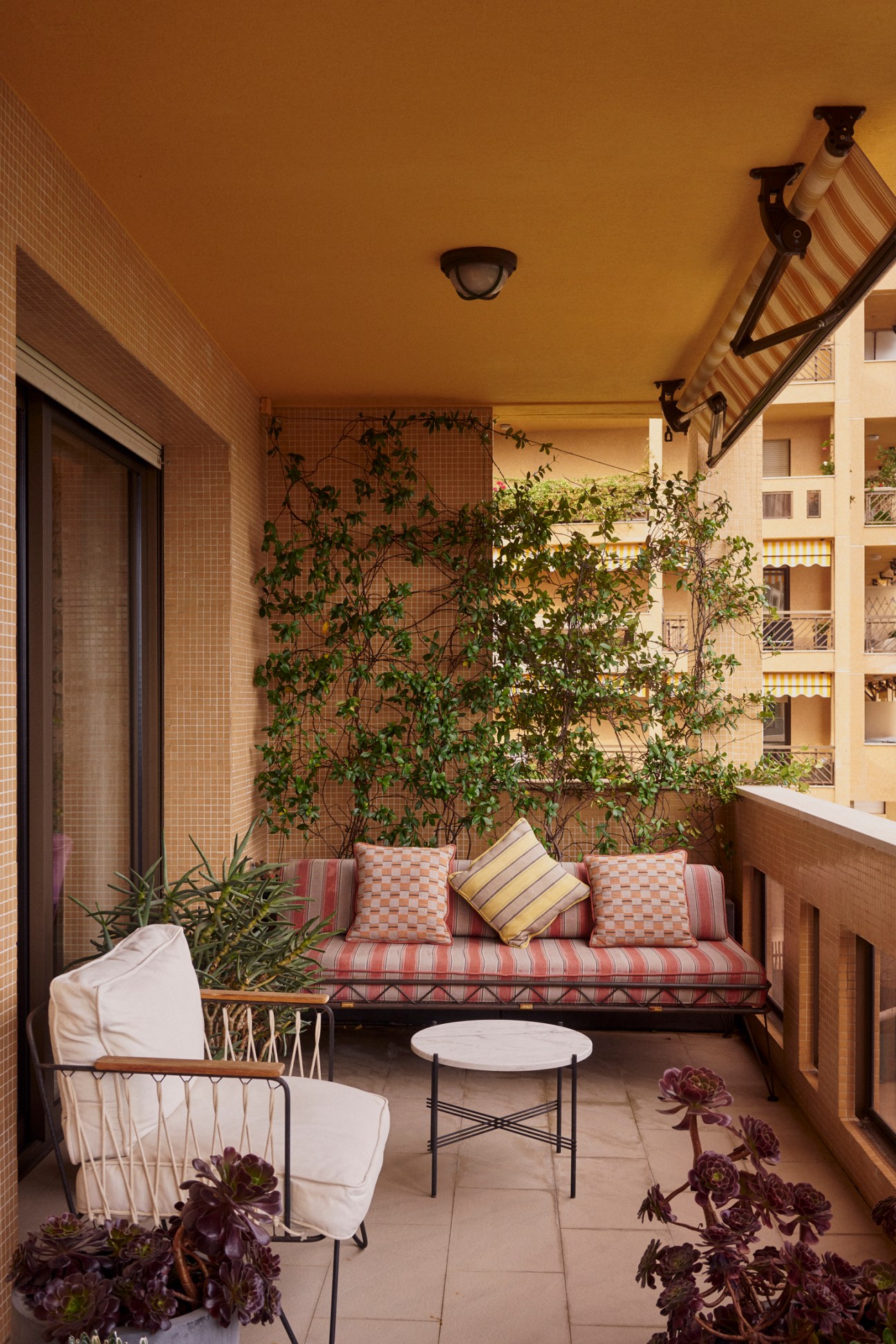 Covered balcony with striped cushioned seating, plants, and small round table overlooking residential buildings