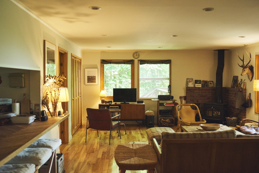 Interior of a sitting room with wooden floors, large windows showing forest views, fireplace, and mixed furnishings including couches and a desk area.