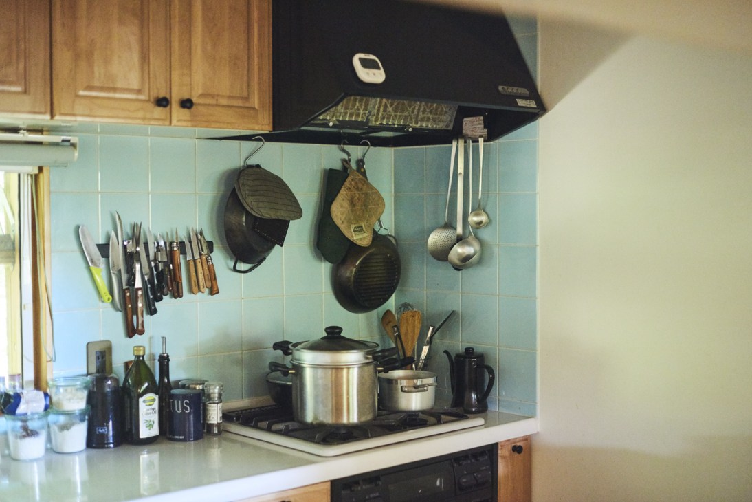 Kitchen with light blue tile backsplash, wooden cabinets, gas stovetop, and cooking utensils and pans hanging on hooks.