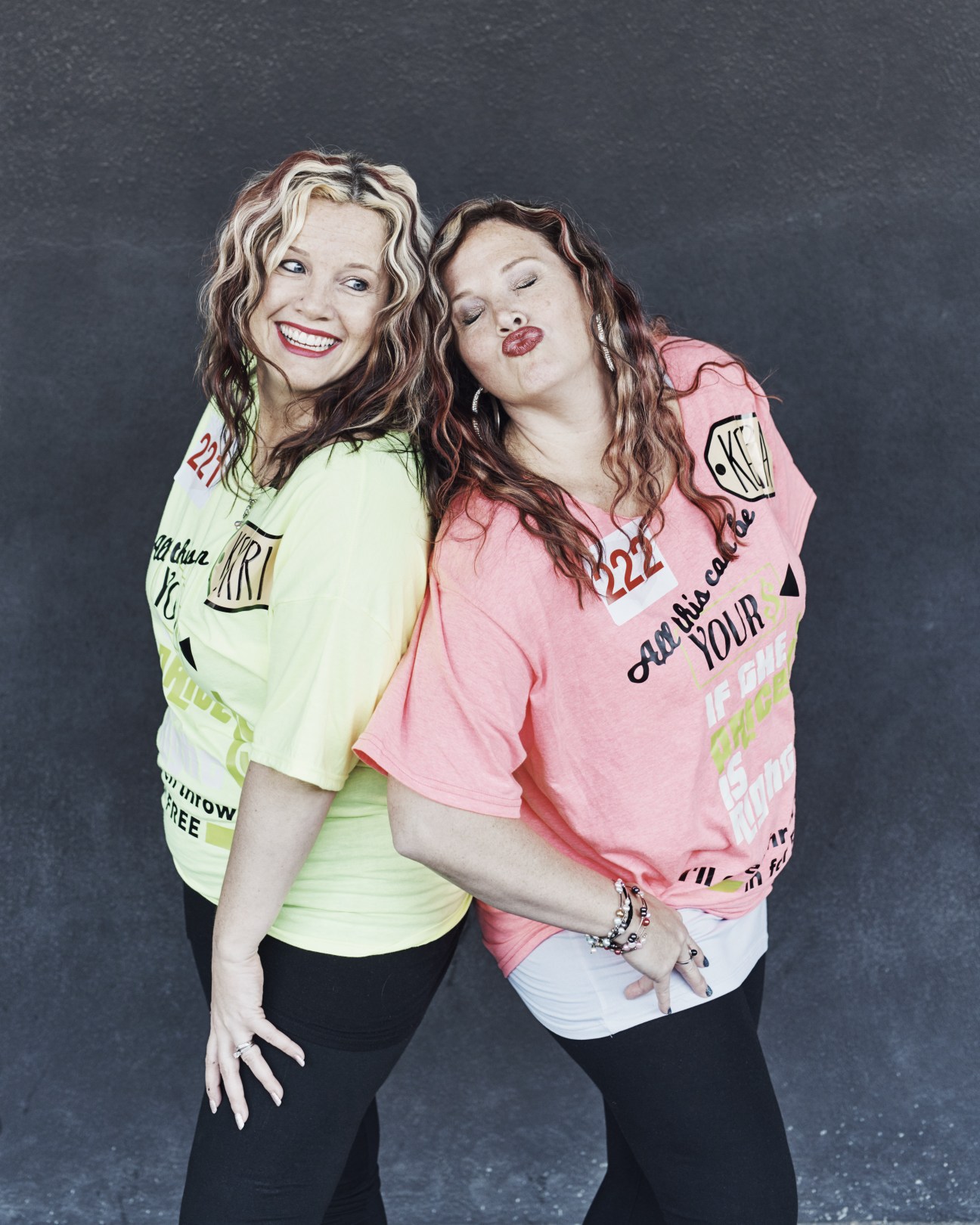 Two women in colorful graphic t-shirts pose together against a dark background.