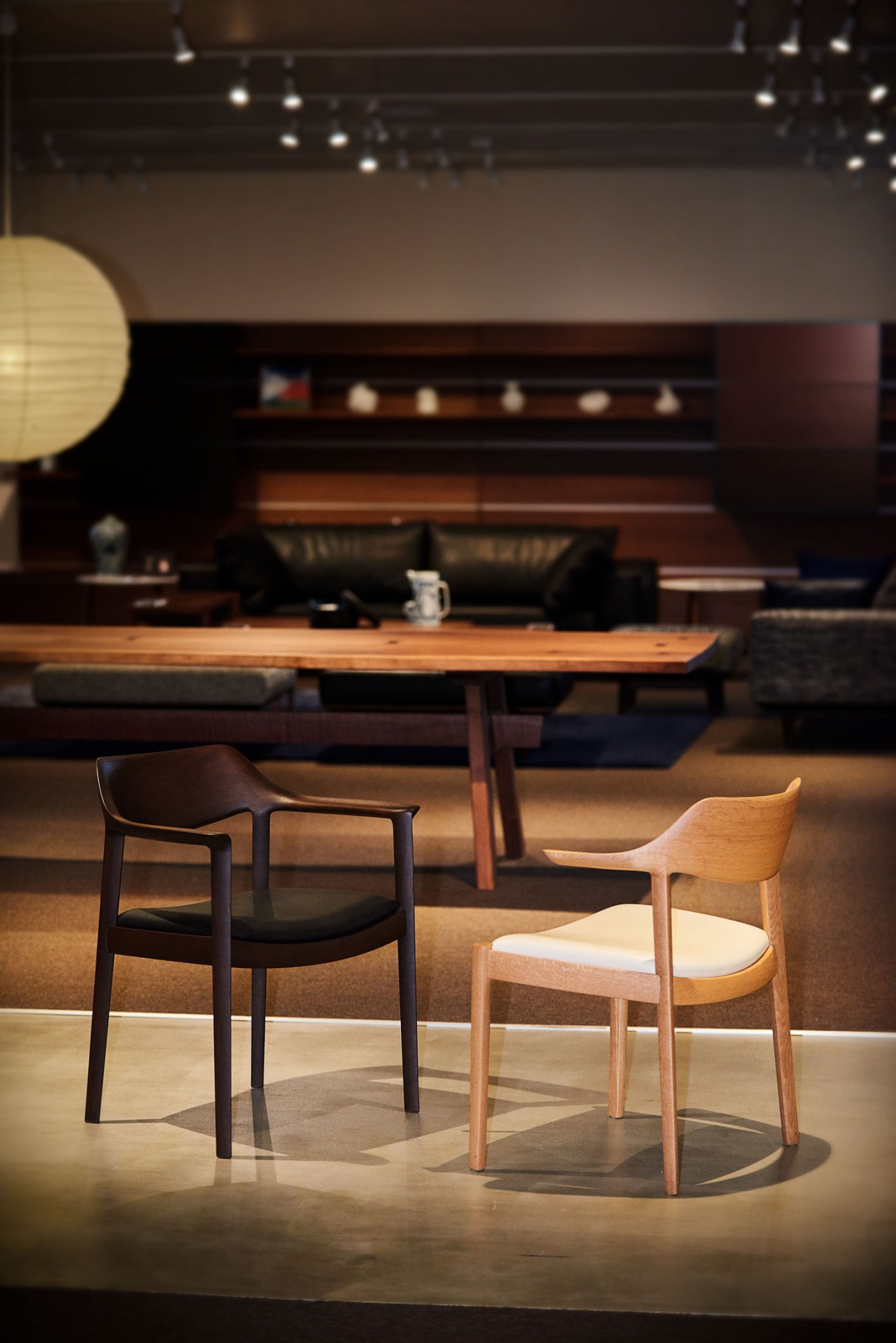 Two wooden chairs displayed in a showroom: one dark chair with black upholstery and one light natural wood chair with cream cushion.