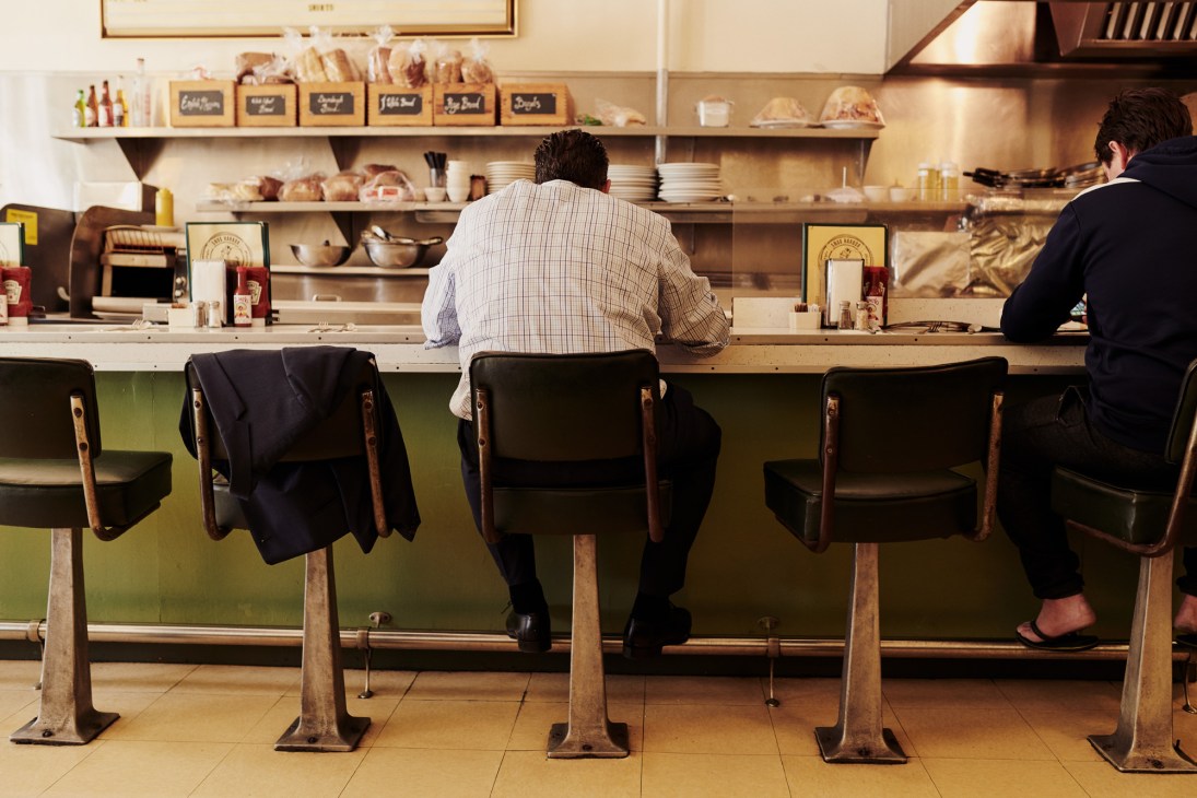 Man seated at a lunch counter with vintage stools at a 1940s-style diner with green paneling and open shelving.
