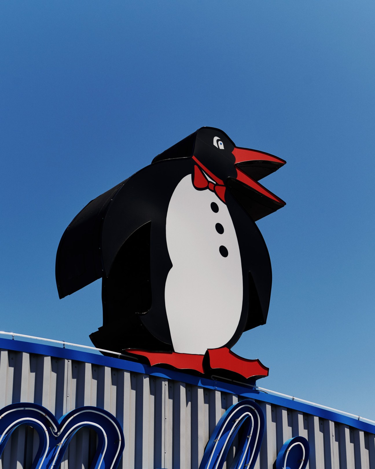 Black penguin mascot with red bow tie and feet on a blue-roofed building against clear sky