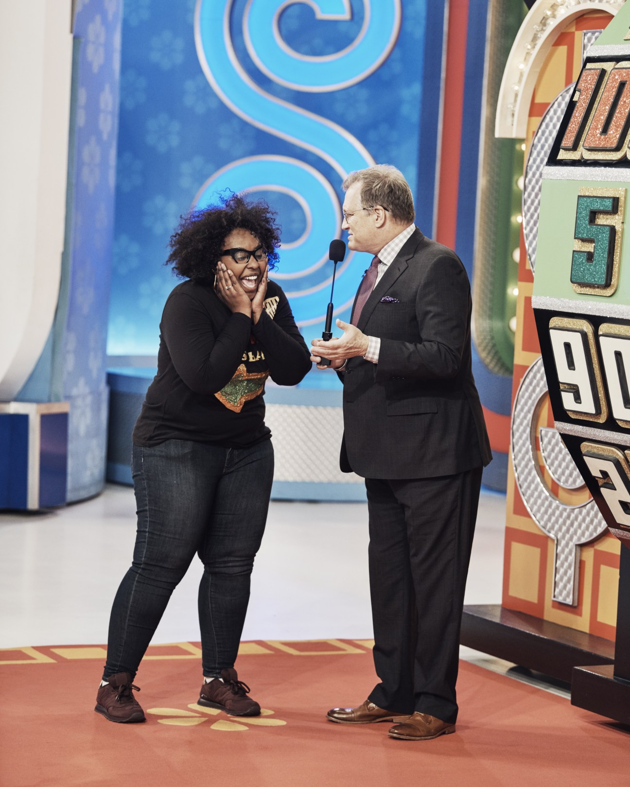 Drew Carey in a dark suit talking to a smiling contestant on The Price is Right stage set.