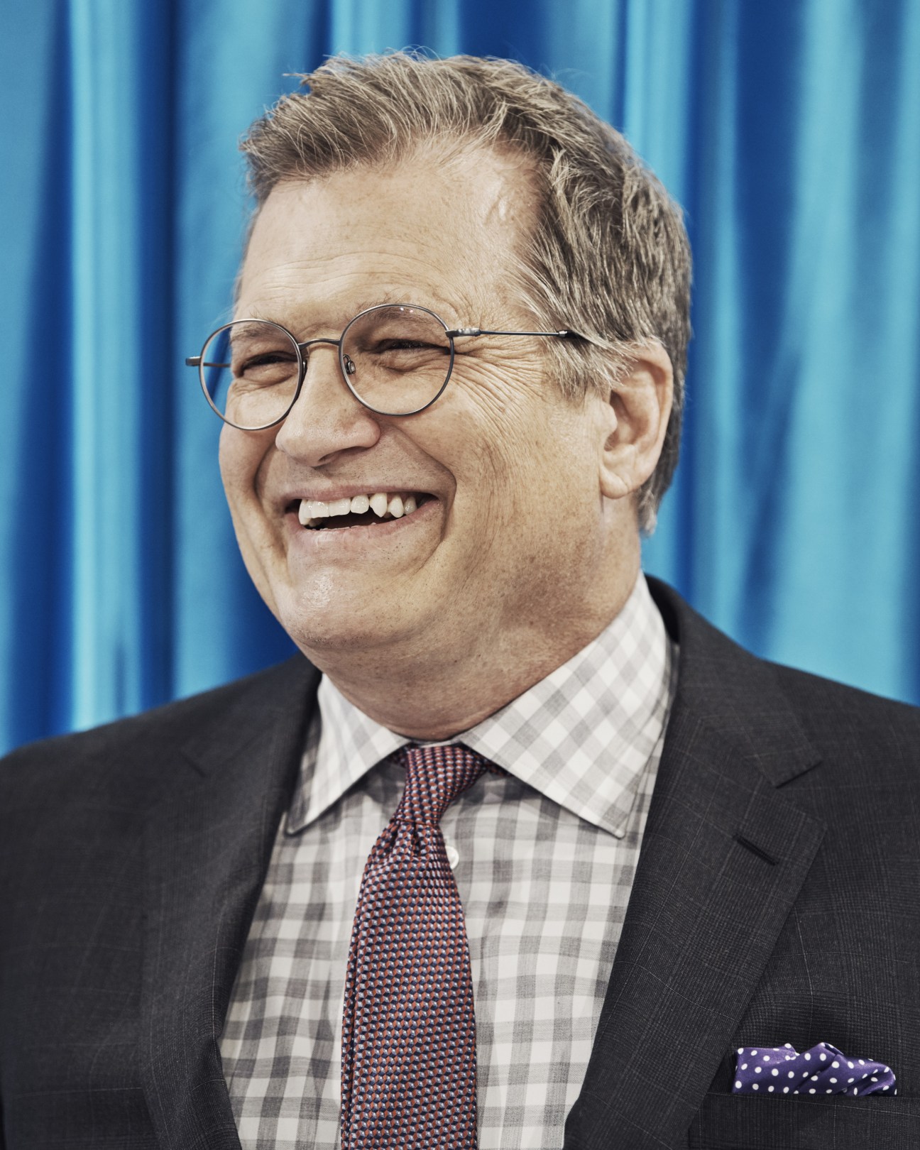Drew Carey smiling in a dark blazer, checkered shirt and patterned tie against a blue curtain backdrop