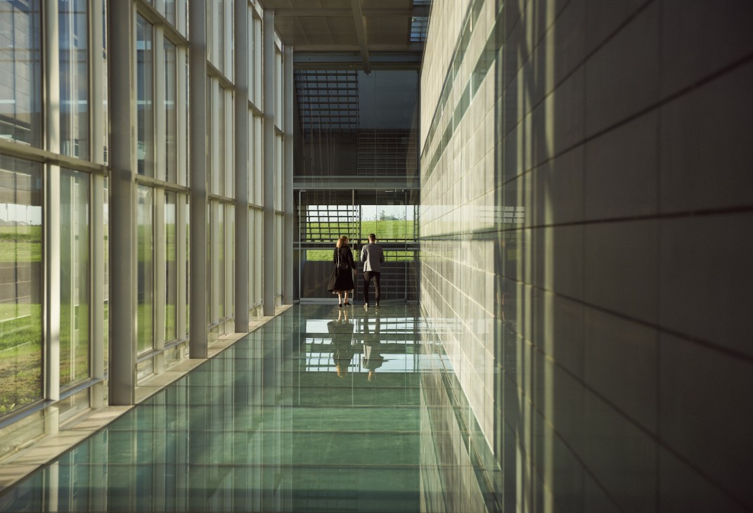 Two people walk through a modern corridor with glass walls and green fields visible outside.