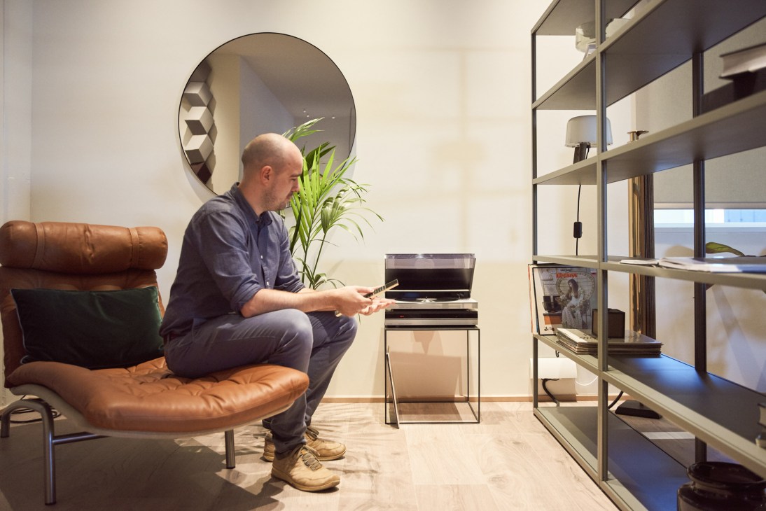 Man sitting on brown leather chair in modern living room with Bang & Olufsen products displayed on shelving.