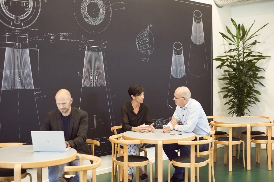 Three employees collaborate at a round table in a modern office with product design blueprints displayed on the wall behind them.