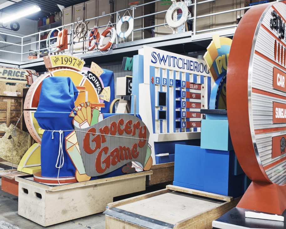 Game show props and set pieces stored in a warehouse, including colorful game boards and vintage carnival-style equipment.