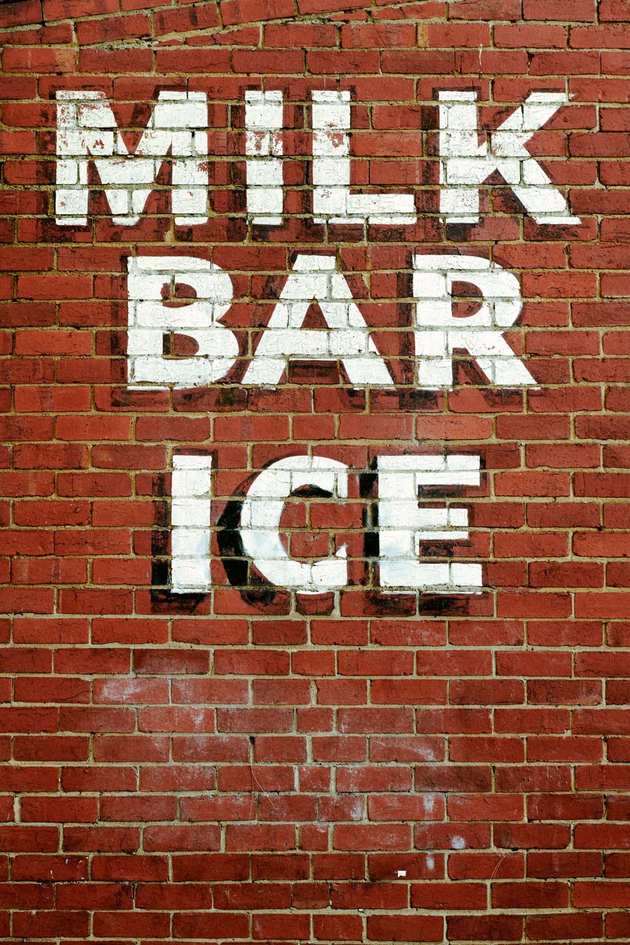 Vintage white painted signage reading 'Milk Bar Ice' on weathered red brick wall.