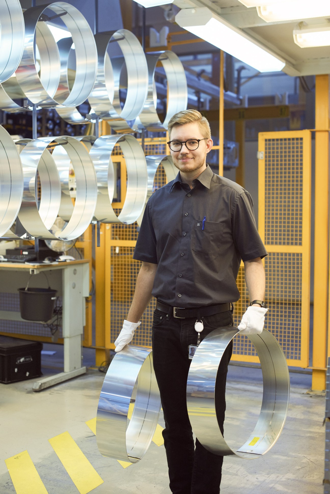 Factory worker holding polished aluminium speaker component in B&O manufacturing facility.