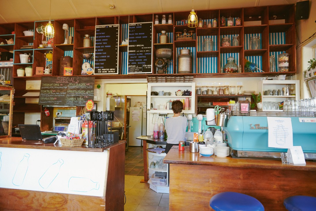 Interior of Cowderoy's Dairy showing wooden counter, shelving with books and merchandise, cone-shaped pendant lights, and a barista at the espresso machine.
