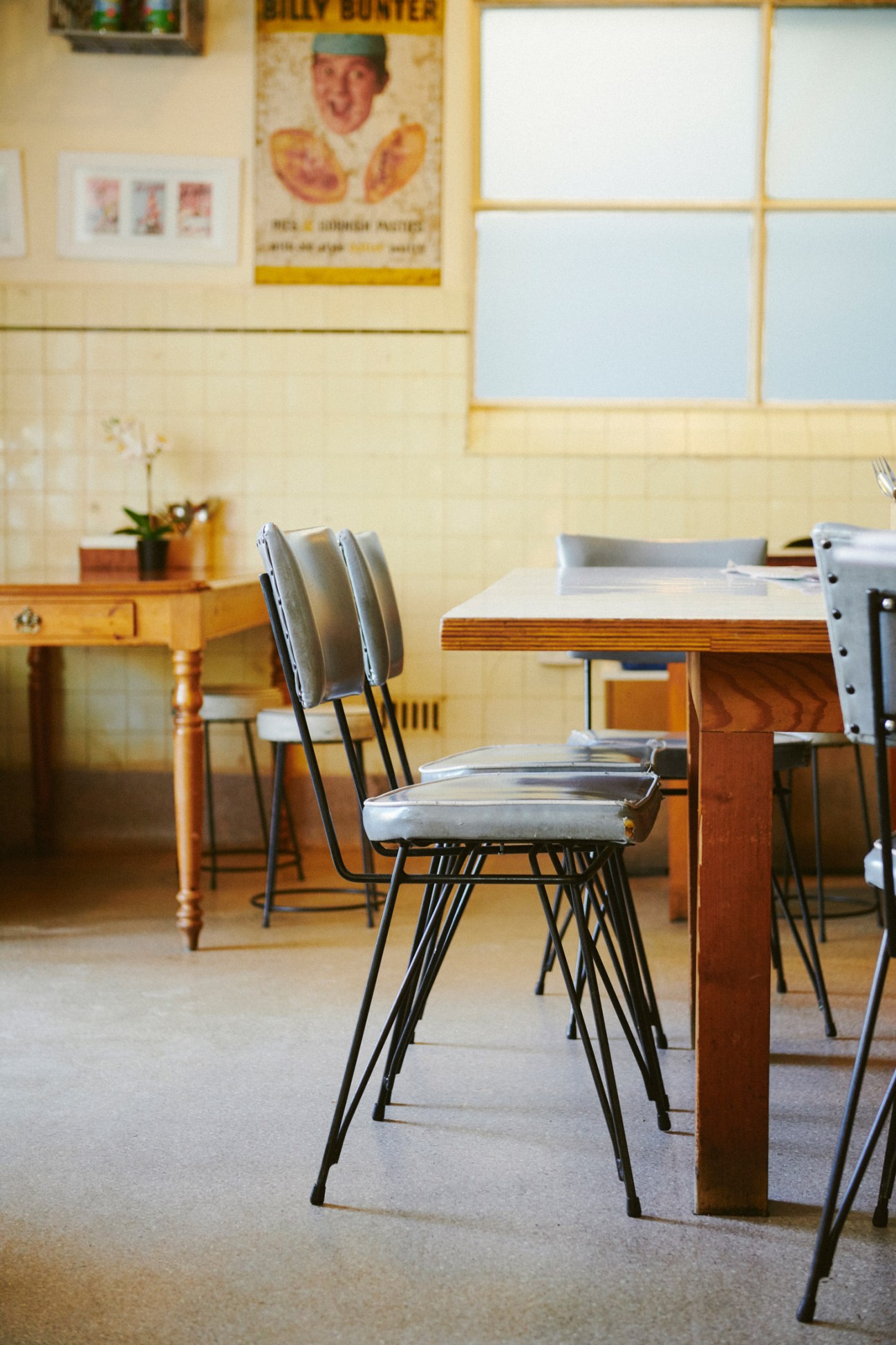 Mid-century metal chairs with gray vinyl seats at a wooden table in a cafe with cream walls and vintage signage.