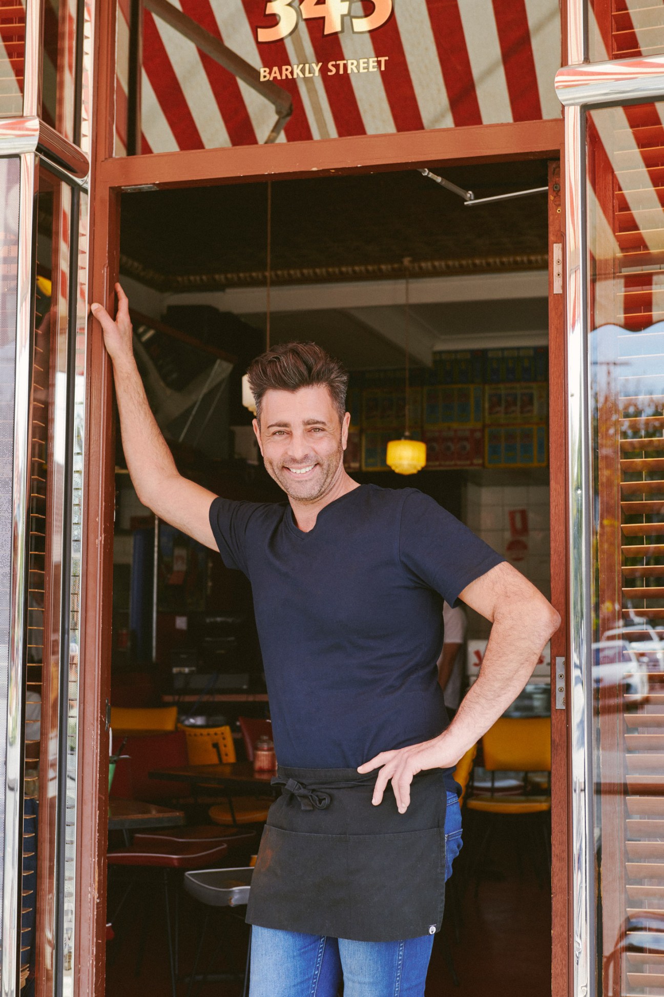 Andrew Serratore stands smiling in the doorway of Jerry's Milk Bar on Barkly Street, with the red and white striped awning visible above.