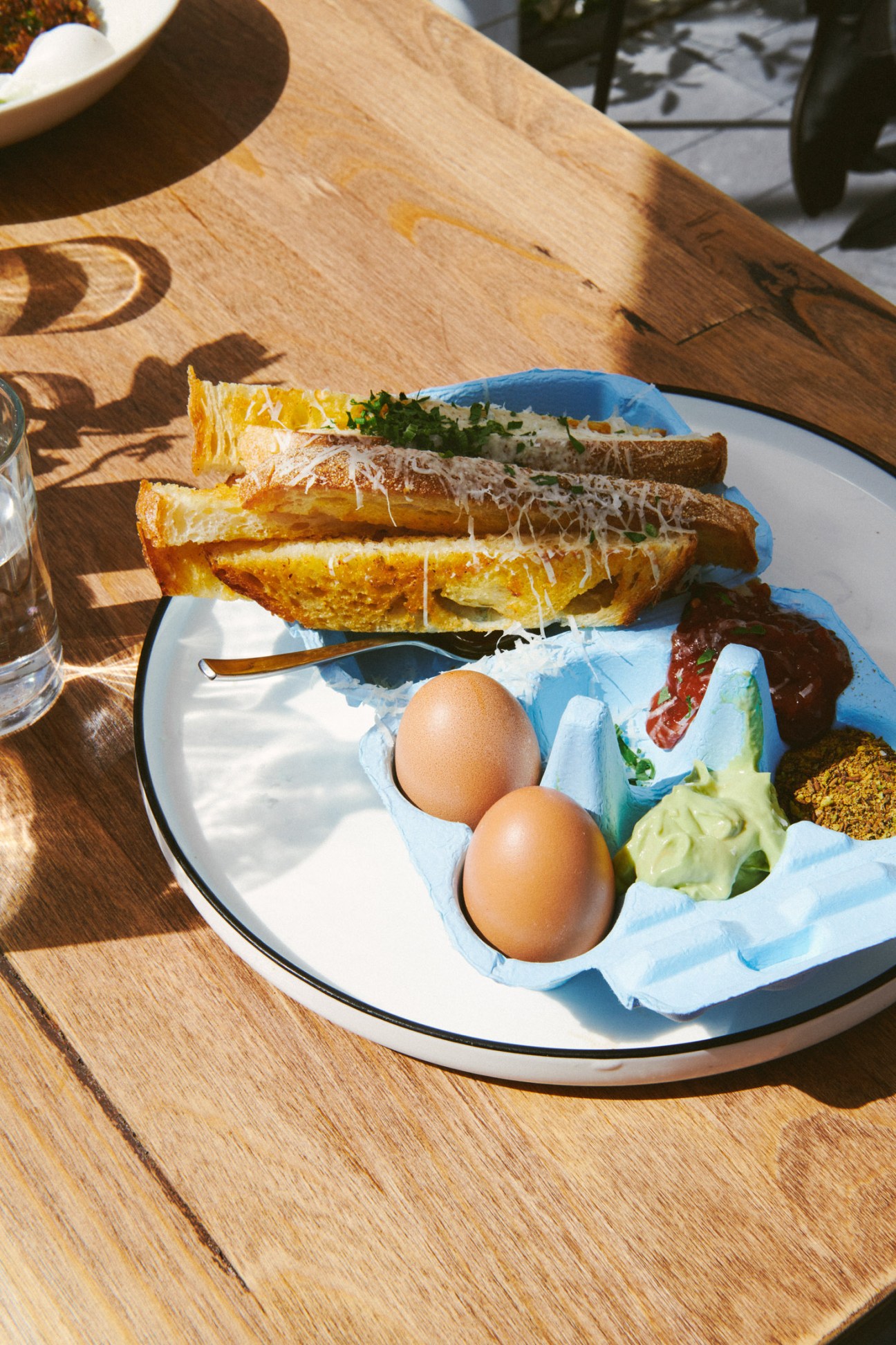 Egg and soldiers breakfast dish with toasted bread strips, two eggs in blue egg cup, and greens on wooden table in sunlight.