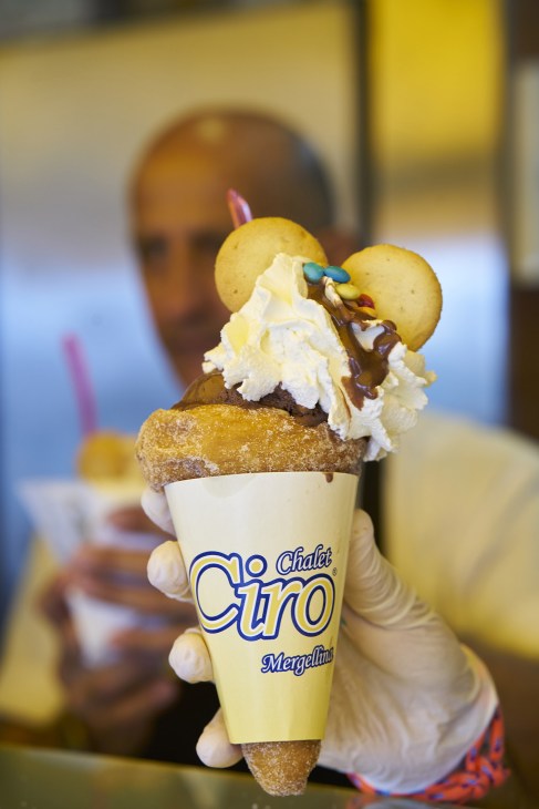 A hand holding a Chalet Ciro gelato cone with whipped cream, chocolate sauce and candy eyes on top.