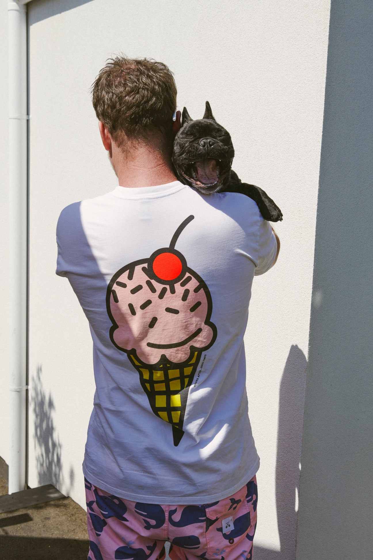 Man wearing ice-cream graphic t-shirt holds a black French bulldog on his shoulder.