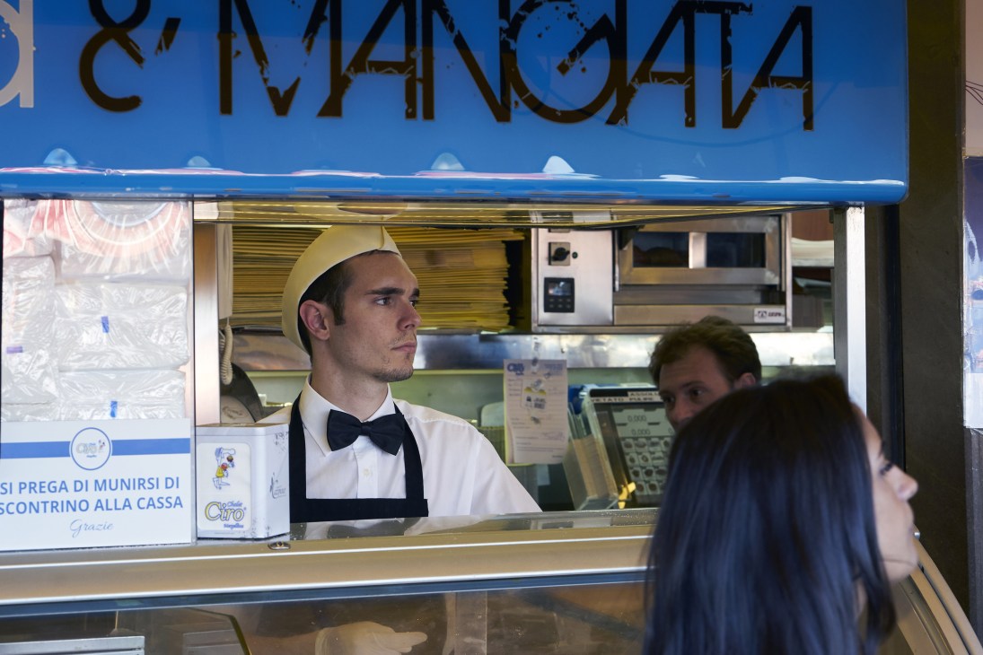 A formal waiter in a yellow hat and black bow tie serves customers at an ice cream shop counter in Naples.