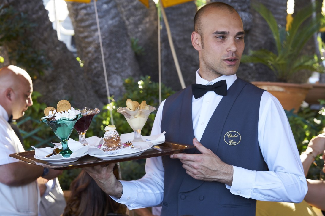 Waiter in formal attire holding a tray of colorful desserts and ice cream at an outdoor restaurant.