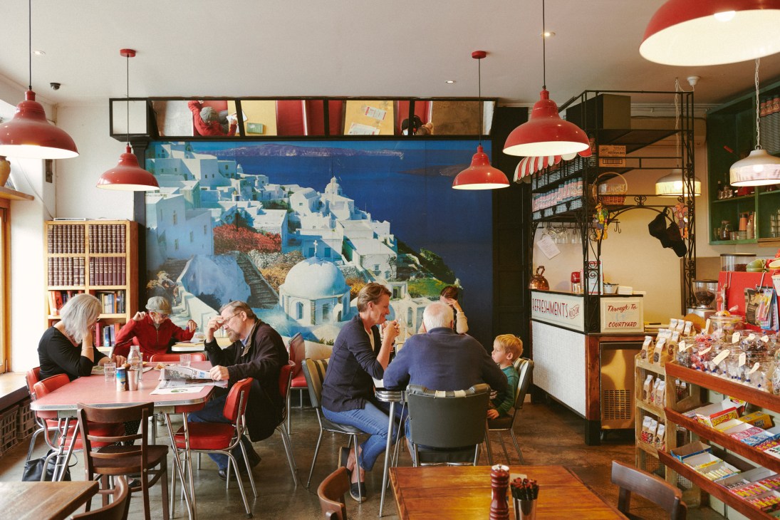 Interior of Rowena Corner Store with customers seated at tables under red pendant lights beneath a Santorini fresco mural.