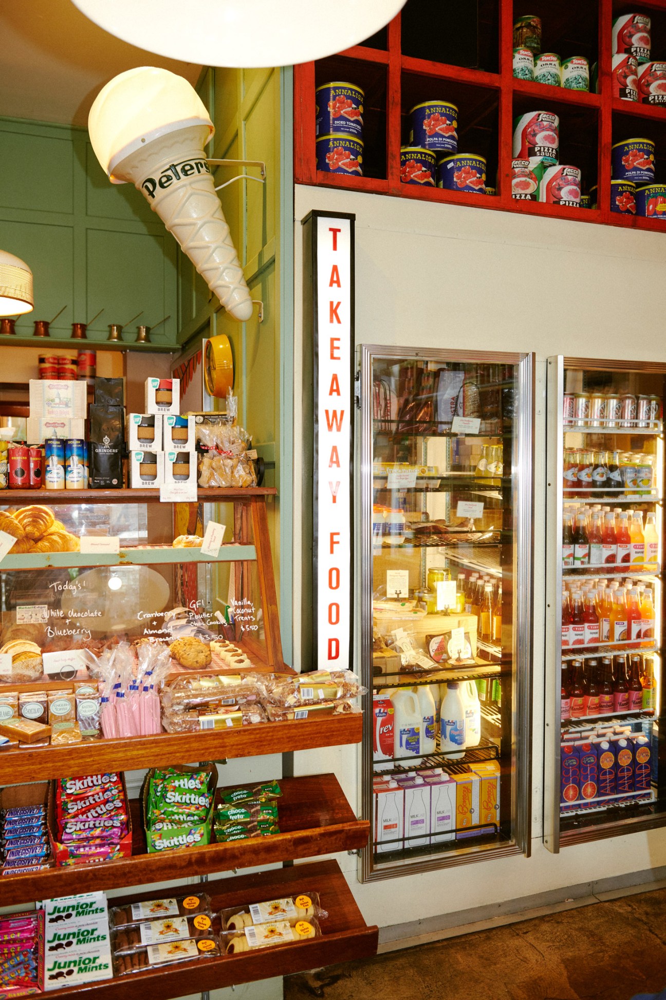 Interior of Rowena Corner Store displaying vintage ice-cream cone light fixture, red shelving with products, refrigerated cases, and candy displays.
