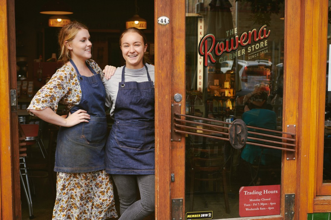 Two staff members smiling outside Rowena Corner Store, a vintage-style milk bar with wooden storefront and red signage.