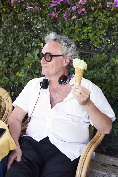 Man with grey hair and glasses holding a vanilla ice cream cone, wearing headphones around his neck and a white shirt, seated against a flowering plant backdrop.