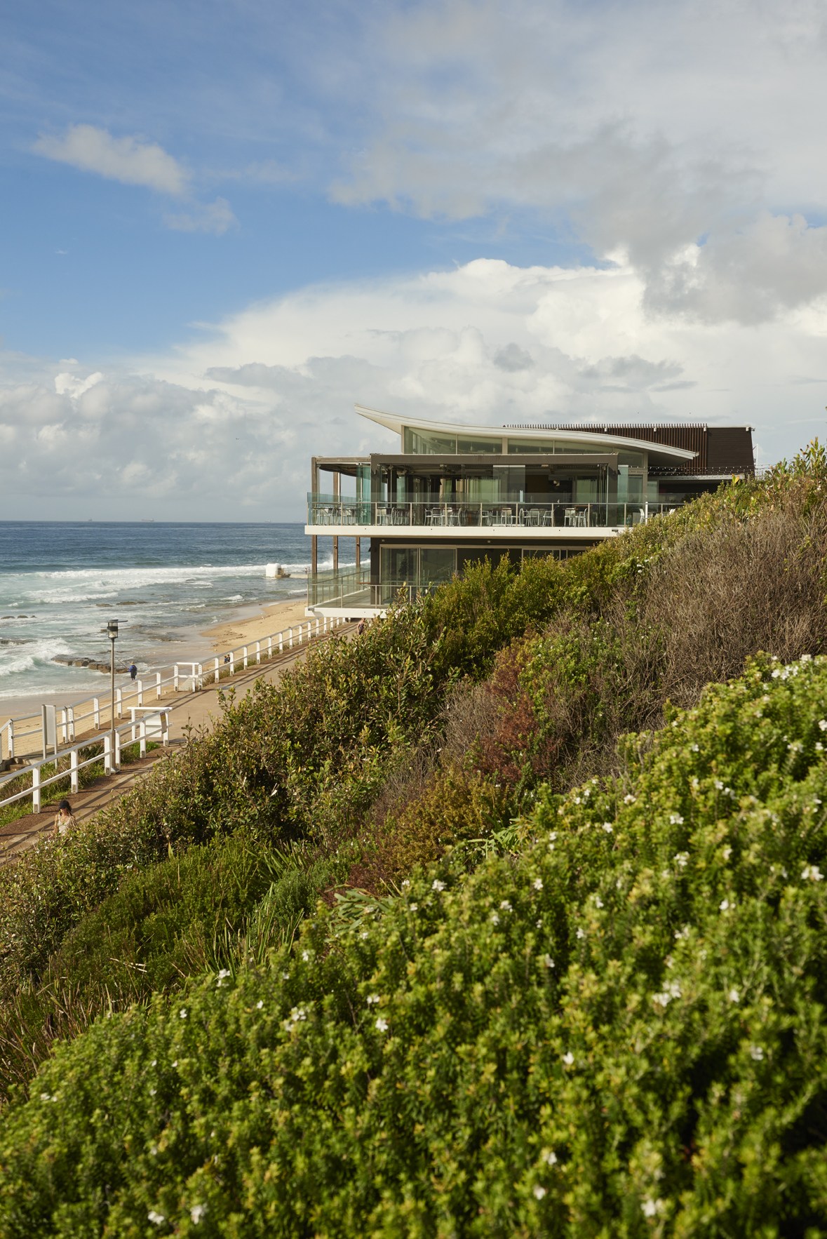 Modern beachfront building with glass panels and angled roof overlooking sandy beach and ocean waves.