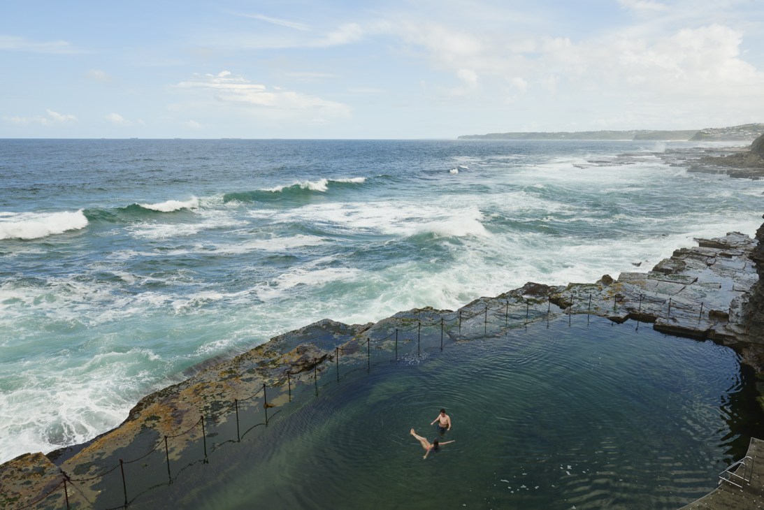 A swimmer in a natural rock pool surrounded by cliffs overlooking the ocean with rolling waves beyond.