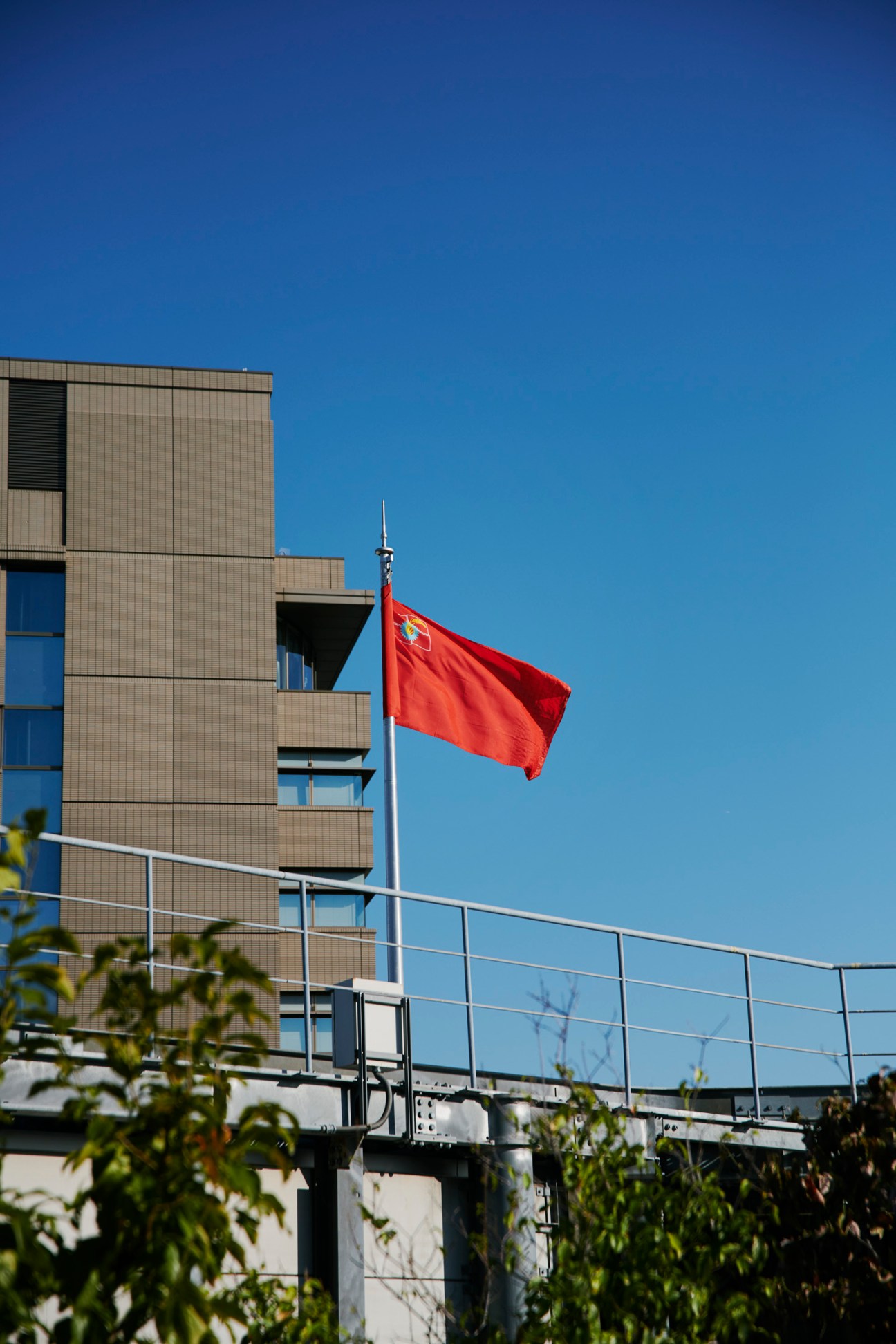 Red flag with emblem flying on flagpole against blue sky next to modern building with metal railings
