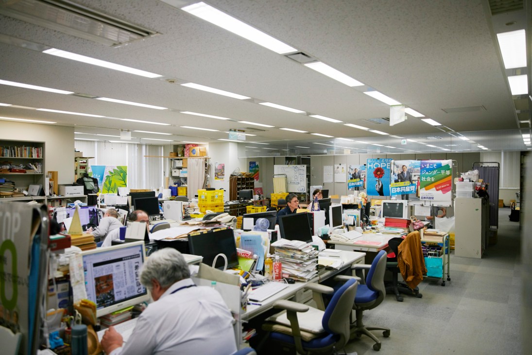 Office workers at cluttered desks with computers and stacks of papers in a fluorescent-lit newsroom environment.
