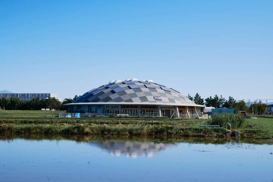 Modern dome-shaped building with checkerboard facade sits in rural farmland beside a reflective water channel