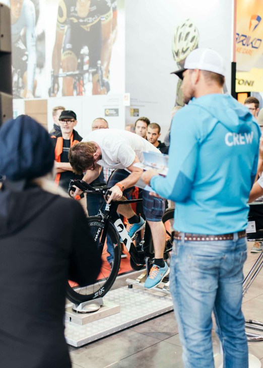 A child sits on a black electric bike at the Eurobike trade fair while a crew member in a blue jacket assists.