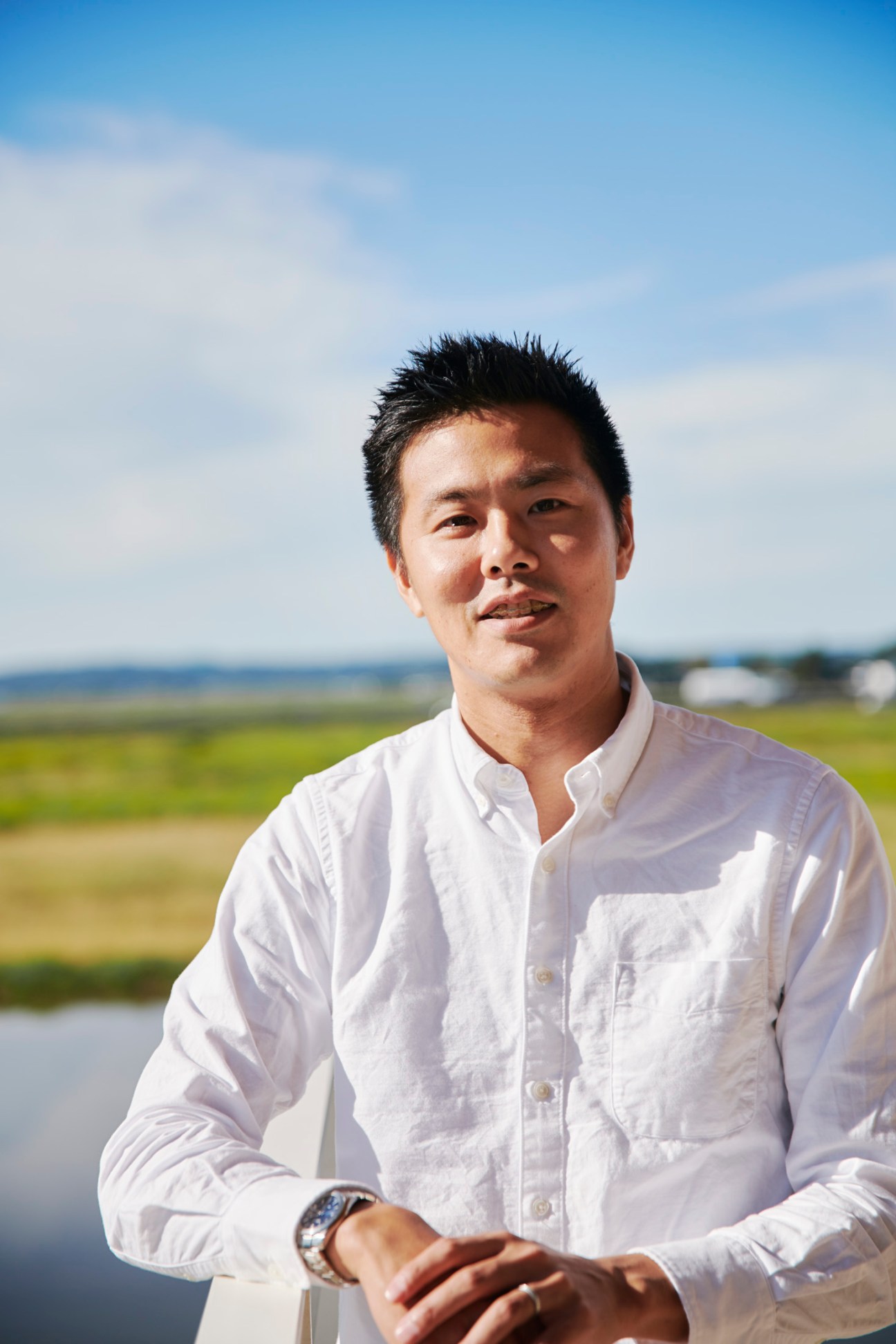 Asian man in white button-down shirt with crossed arms stands outdoors against rural landscape with blue sky