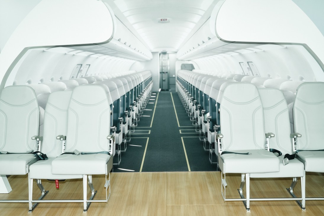 Interior view of an aircraft cabin with rows of white seats and a central aisle extending toward the exit.