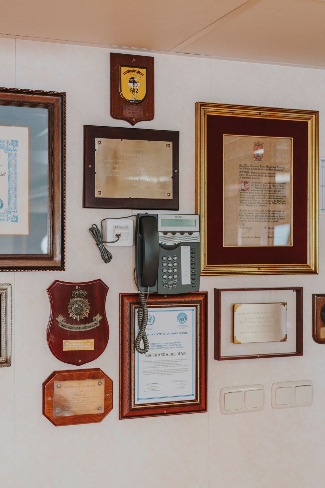 Wall display of framed certificates, awards, and plaques recognizing the ship's humanitarian work, with a mounted wooden emblem and vintage telephone.