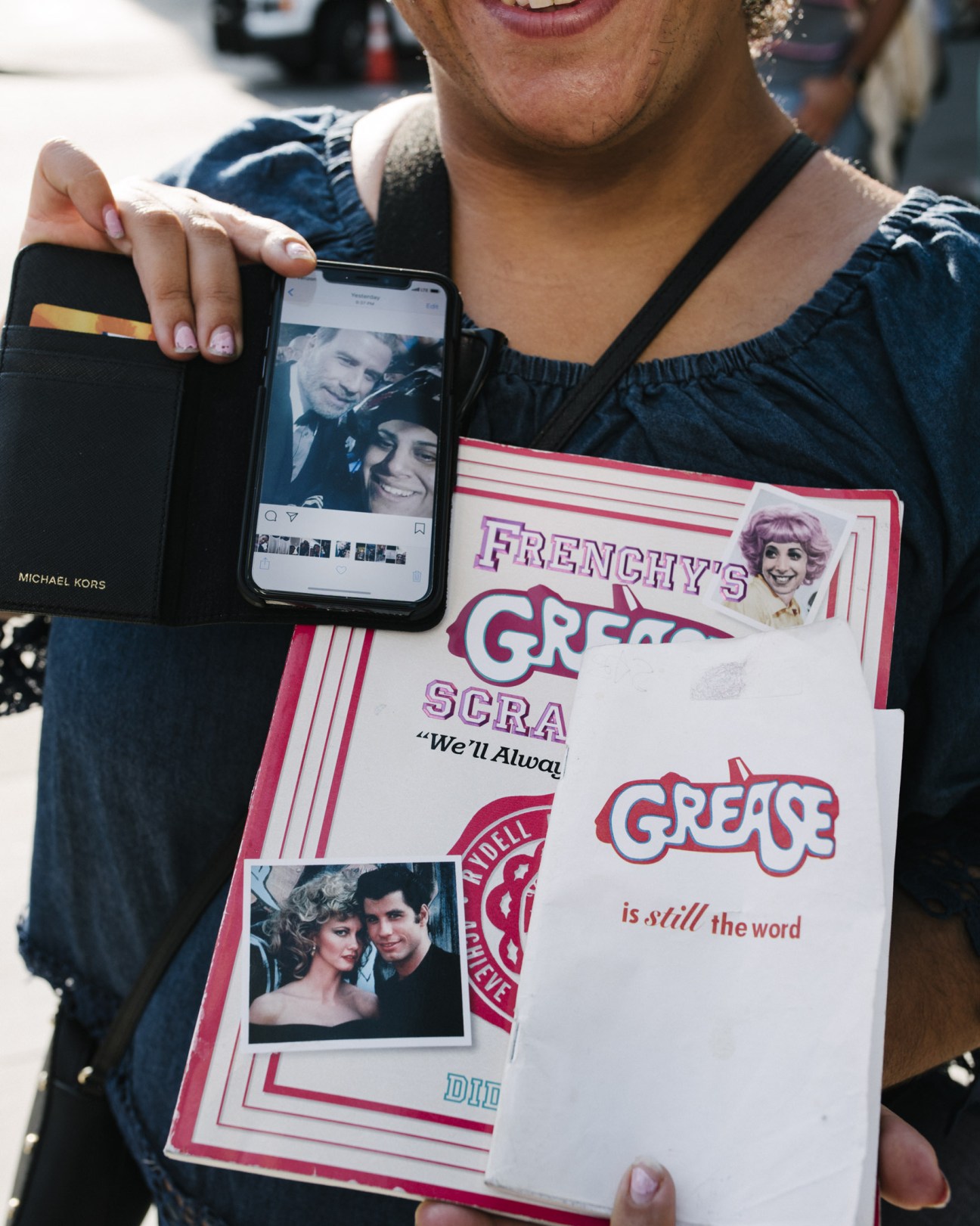 Person holding a smartphone displaying a selfie and a Grease movie poster with merchandise.