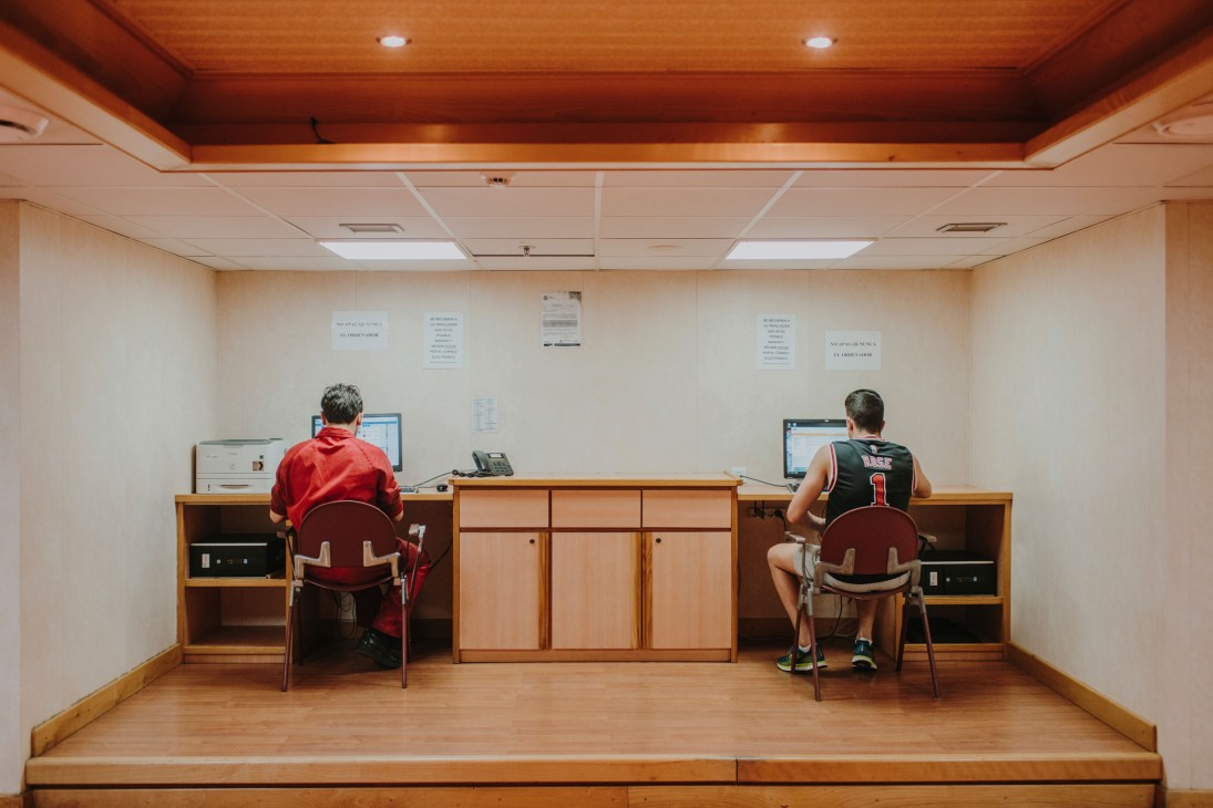 Two crew members working at computer terminals on opposite sides of a shared wooden desk in a ship's office with beige walls and wooden ceiling.