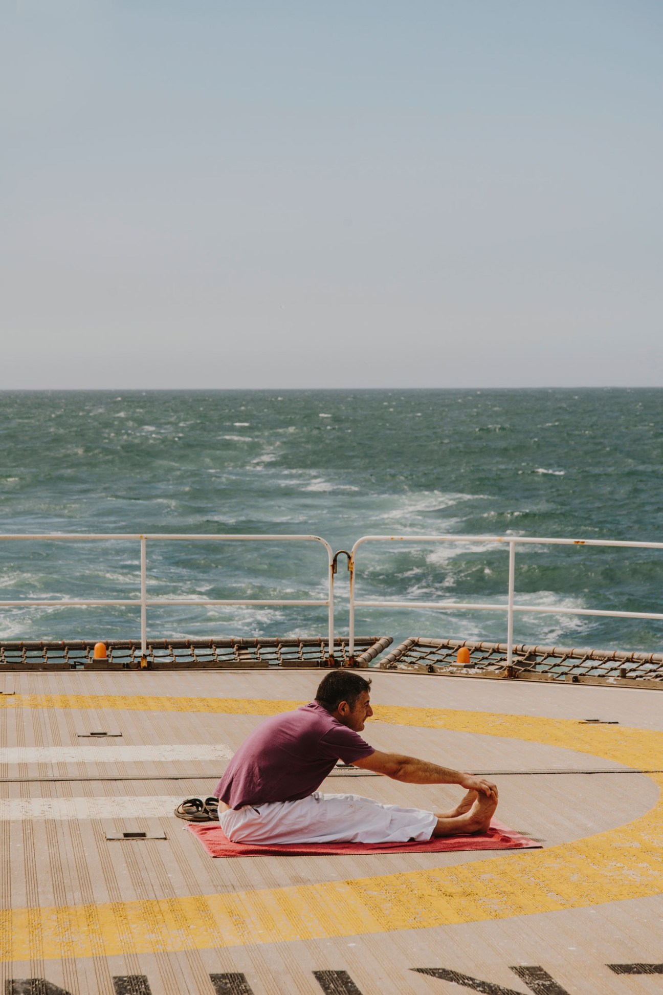 Man in purple shirt performing seated forward bend yoga on red mat overlooking ocean from ship deck.