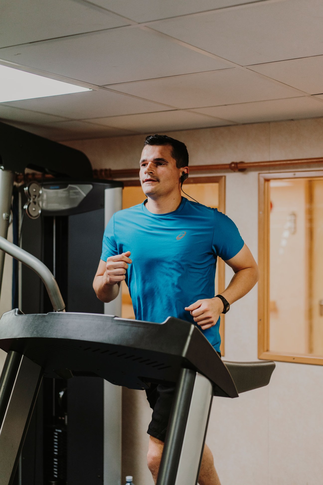 Man in blue athletic shirt running on treadmill in ship's gym with drop ceiling and wooden doors.
