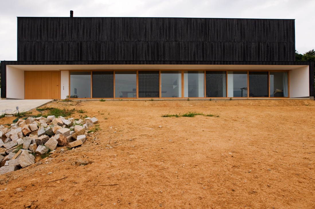 Modern house with burnt cedar cladding, white walls, sliding glass windows, and wooden door on a bare hillside.
