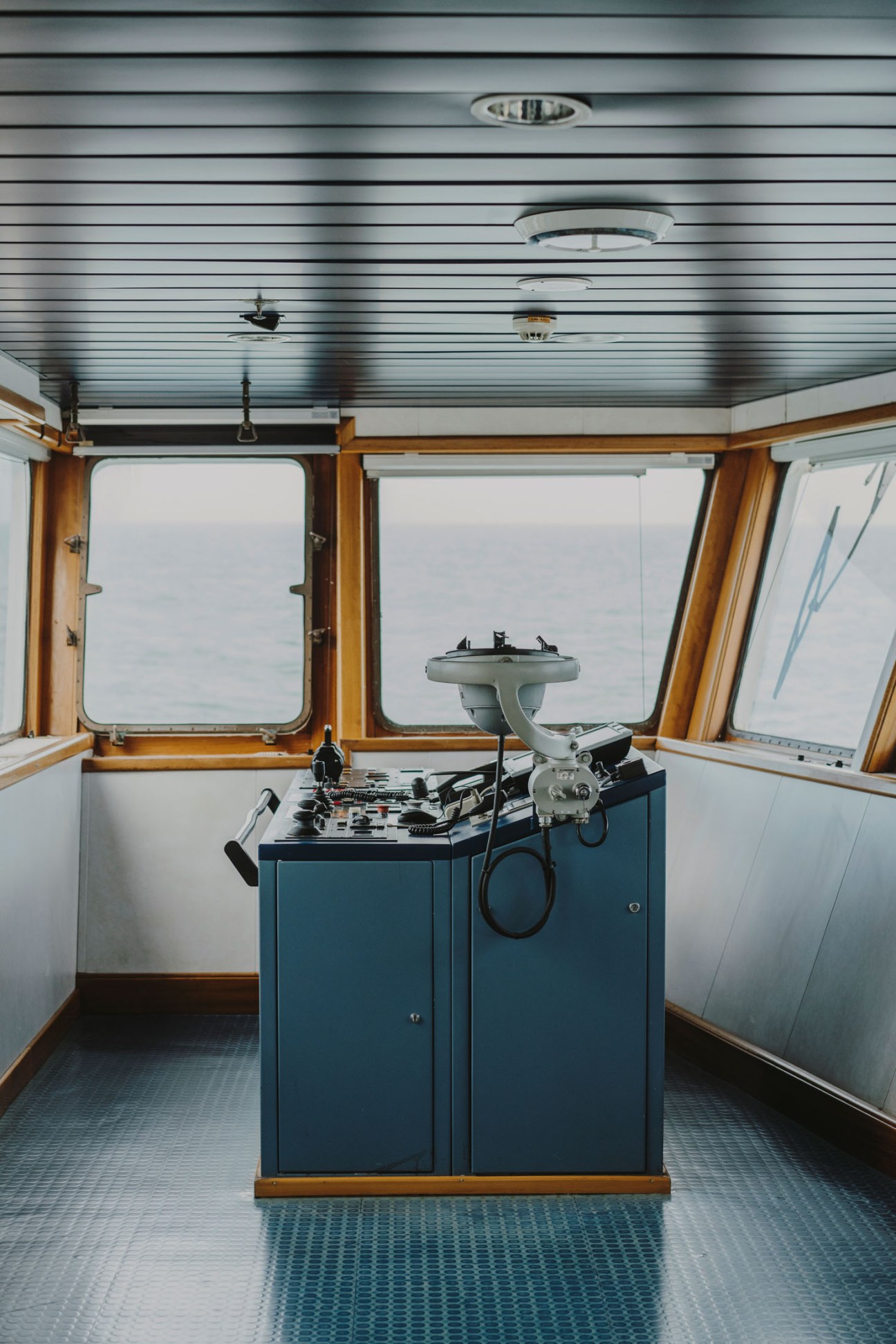 Ship's bridge control panel with navigation equipment mounted on a blue cabinet beneath wooden-framed windows overlooking the ocean.
