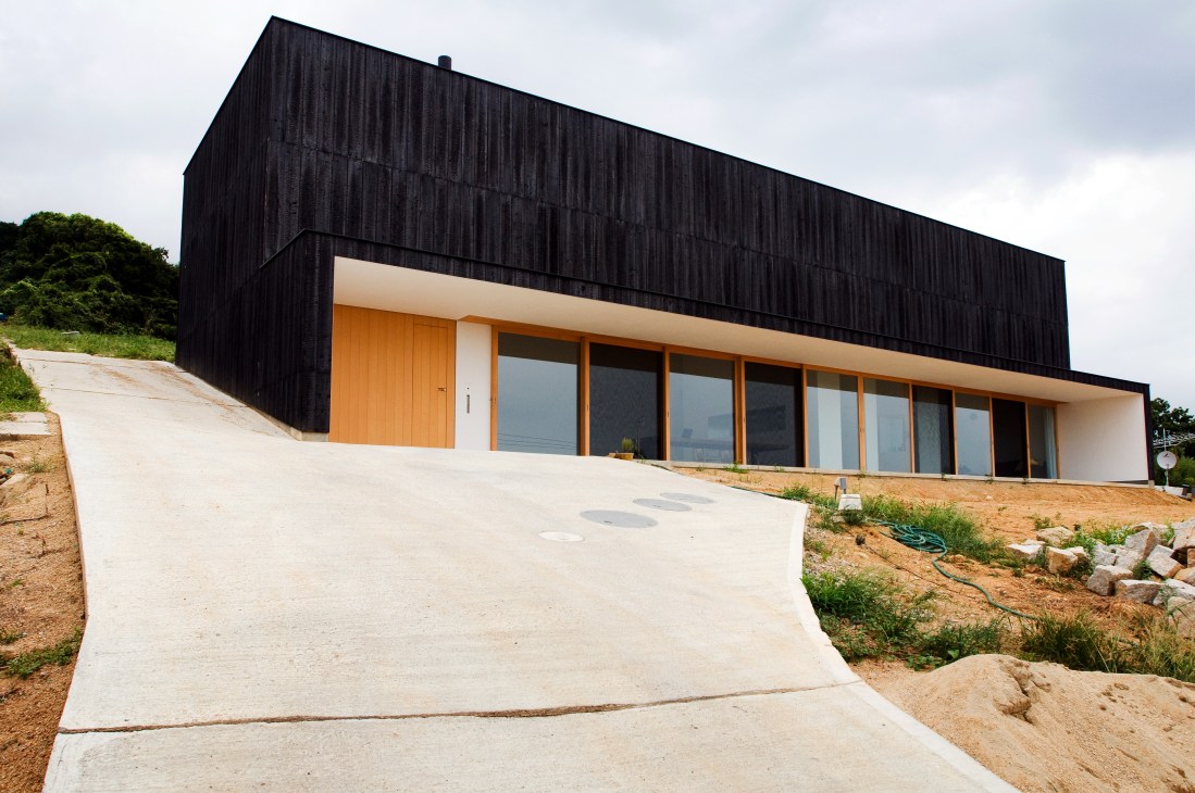 Modern house with black burnt cedar cladding, white facade, orange door, and large windows overlooking countryside.