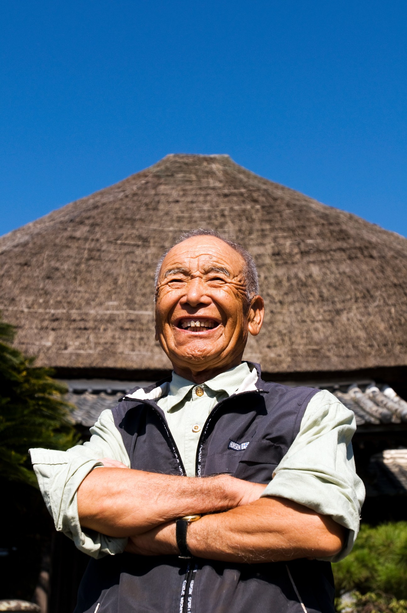 Master thatcher Kazuyoshi Koga smiling with arms crossed in front of a traditional Japanese thatched-roof farmhouse.