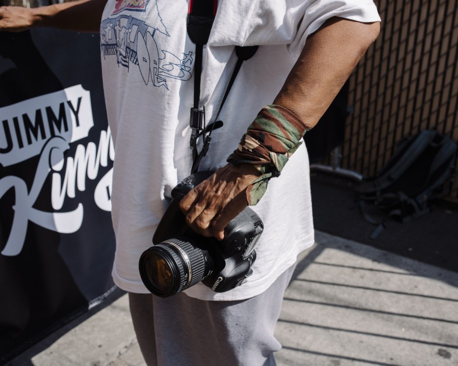 A paparazzo holds a professional camera with a long lens, wearing fabric wraps on their wrist.